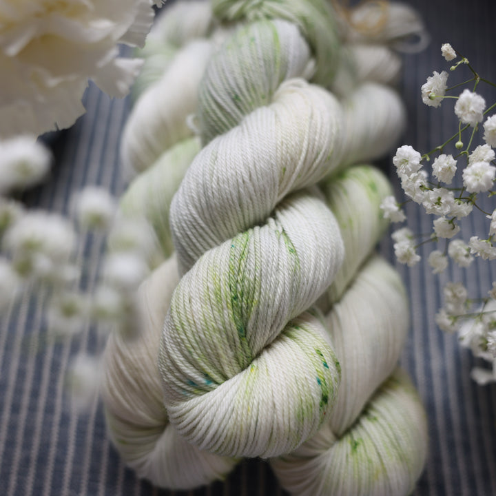 DK yarn colored a white and very light green with some darker green speckles. These skeins are on top of a faded navy blue and white striped fabric with white flowers like roses and babies breath around the yarn.
