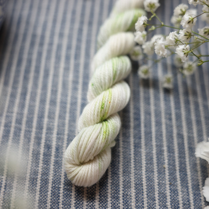 DK mini yarn colored a white and very light green with some darker green speckles. This skein is on top of a faded navy blue and white striped fabric with white flowers like roses and babies breath around the yarn.