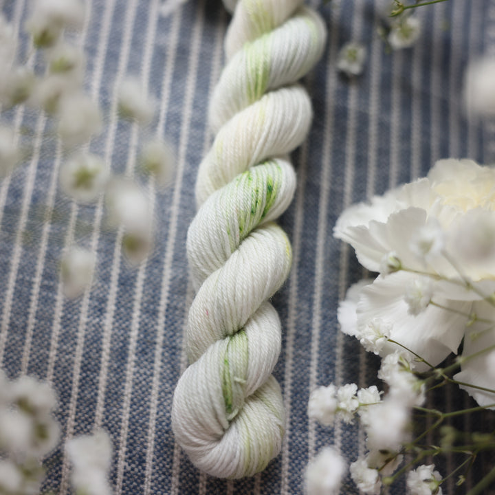 Fingering mini yarn colored a white and very light green with some darker green speckles. These skeins are on top of a faded navy blue and white striped fabric with white flowers like roses and babies breath around the yarn.