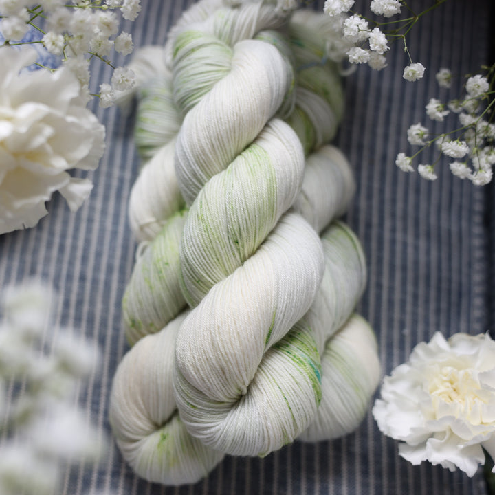 Fingering yarn colored a white and very light green with some darker green speckles. These skeins are on top of a faded navy blue and white striped fabric with white flowers like roses and babies breath around the yarn.