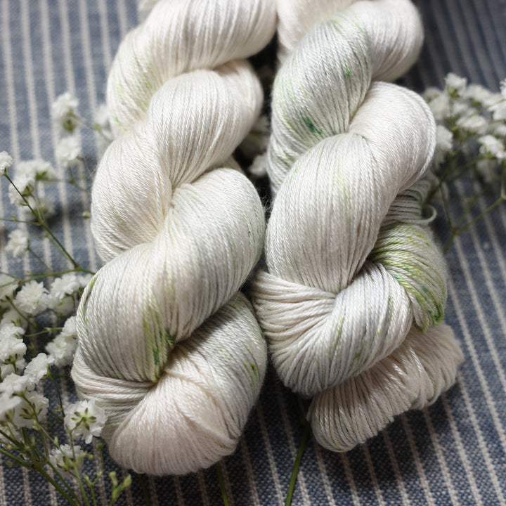 Silk fingering yarn colored a white and very light green with some darker green speckles. These skeins are on top of a faded navy blue and white striped fabric with white flowers like roses and babies breath around the yarn.