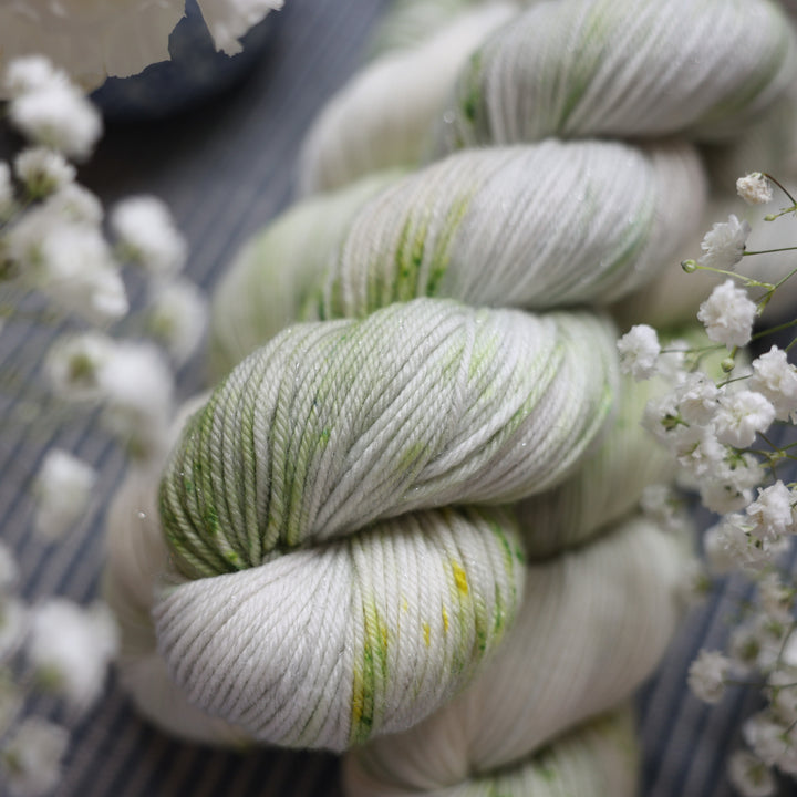 Sparkle DK yarn colored a white and very light green with some darker green speckles. These skeins are on top of a faded navy blue and white striped fabric with white flowers like roses and babies breath around the yarn.
