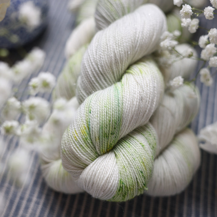Sparkle fingering yarn colored a white and very light green with some darker green speckles. These skeins are on top of a faded navy blue and white striped fabric with white flowers like roses and babies breath around the yarn.