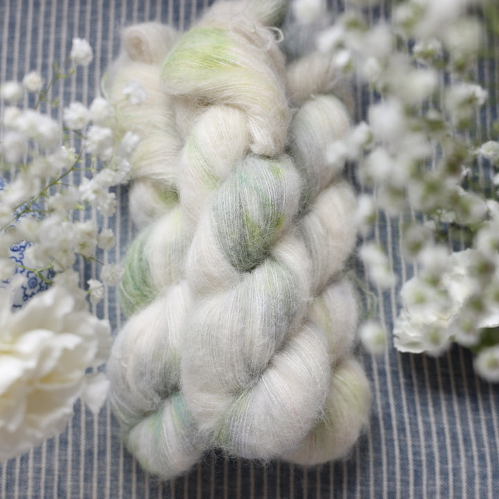 Suri yarn colored a white and very light green with some darker green speckles. These skeins are on top of a faded navy blue and white striped fabric with white flowers like roses and babies breath around the yarn.