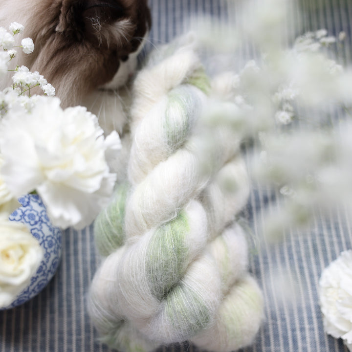Suri yarn colored a white and very light green with some darker green speckles. These skeins are on top of a faded navy blue and white striped fabric with white flowers like roses and babies breath around the yarn. a rag doll cat named Tina is in the corner.