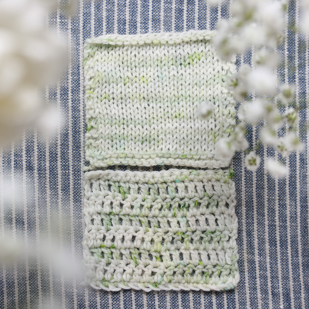 Knit and crochet swatches colored a white and very light green with some darker green speckles. These swatches are on top of a faded navy blue and white striped fabric with white flowers like roses and babies breath around the yarn.