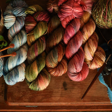 Four DK sparkle-weight skeins from left to right are colored a vibrant blue with pink and green highlights and shifting blue-to-pink speckles, a fawn and cream blend with blush washes and brown speckles,   and a leather brown with mauve, blue, and red speckles, a deep plum with hints of crimson and cool-toned purple, a warm green blend with touches of mauve, brown, and blue,.  In the corner there is a blue ribbon and a leaf hovering over the skeins. All sitting on top of a wooden table.