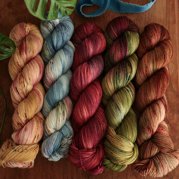 Four fingering-weight skeins from left to right are colored a fawn and cream blend with blush washes and brown speckles, a vibrant blue with pink and green highlights and shifting blue-to-pink speckles, a deep plum with hints of crimson and cool-toned purple, a warm green blend with touches of mauve, brown, and blue, and a leather brown with mauve, blue, and red speckles.  In the corner there is a blue ribbon and a leaf hovering over the skeins. All sitting on top of a wooden table.