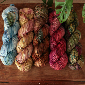 Four DK weight skeins from left to right are colored a vibrant blue with pink and green highlights and shifting blue-to-pink speckles, a fawn and cream blend with blush washes and brown speckles,   and a leather brown with mauve, blue, and red speckles, a deep plum with hints of crimson and cool-toned purple, a warm green blend with touches of mauve, brown, and blue,.  In the corner there is a blue ribbon and a leaf hovering over the skeins. All sitting on top of a wooden table.