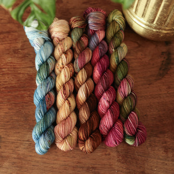 Four fingering Mini-weight skeins from left to right are colored a vibrant blue with pink and green highlights and shifting blue-to-pink speckles, a fawn and cream blend with blush washes and brown speckles,   and a leather brown with mauve, blue, and red speckles, a deep plum with hints of crimson and cool-toned purple, a warm green blend with touches of mauve, brown, and blue,.  In the corner there is a blue ribbon and a leaf hovering over the skeins. All sitting on top of a wooden table.