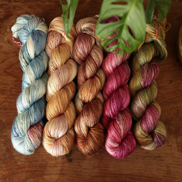 Four silk-weight skeins from left to right are colored a vibrant blue with pink and green highlights and shifting blue-to-pink speckles, a fawn and cream blend with blush washes and brown speckles,   and a leather brown with mauve, blue, and red speckles, a deep plum with hints of crimson and cool-toned purple, a warm green blend with touches of mauve, brown, and blue,.  In the corner there is a blue ribbon and a leaf hovering over the skeins. All sitting on top of a wooden table.