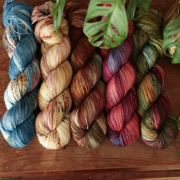 Four sparkle-weight skeins from left to right are colored a vibrant blue with pink and green highlights and shifting blue-to-pink speckles, a fawn and cream blend with blush washes and brown speckles,   and a leather brown with mauve, blue, and red speckles, a deep plum with hints of crimson and cool-toned purple, a warm green blend with touches of mauve, brown, and blue,.  In the corner there is a blue ribbon and a leaf hovering over the skeins. All sitting on top of a wooden table.