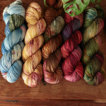Four sport-weight skeins from left to right are colored a vibrant blue with pink and green highlights and shifting blue-to-pink speckles, a fawn and cream blend with blush washes and brown speckles,   and a leather brown with mauve, blue, and red speckles, a deep plum with hints of crimson and cool-toned purple, a warm green blend with touches of mauve, brown, and blue,.  In the corner there is a blue ribbon and a leaf hovering over the skeins. All sitting on top of a wooden table.