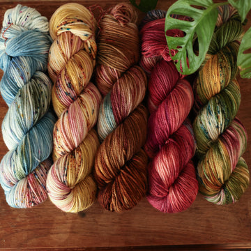 Four worsted-weight skeins from left to right are colored a vibrant blue with pink and green highlights and shifting blue-to-pink speckles, a fawn and cream blend with blush washes and brown speckles,   and a leather brown with mauve, blue, and red speckles, a deep plum with hints of crimson and cool-toned purple, a warm green blend with touches of mauve, brown, and blue,.  In the corner there is a blue ribbon and a leaf hovering over the skeins. All sitting on top of a wooden table.