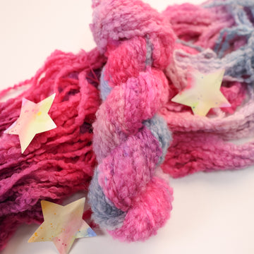 A fluffy yarn colored with different shades of pink, and a grey/blue on top of un unwound version of that yarn, and two multicolored stars next to the yarn.