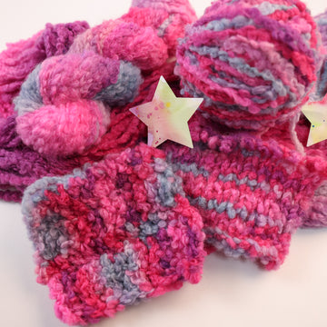 A fluffy yarn colored with different shades of pink, and a grey/blue on top of un unwound version of that yarn, and two multicolored stars next to the yarn, with a knit swatch and crochet swatch of that yarn.