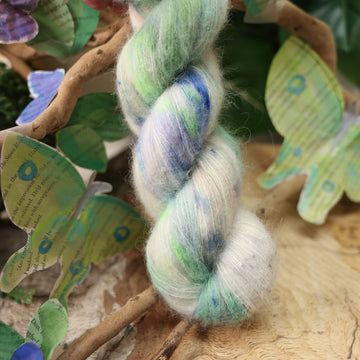 A light blue, teal, green, and white colored soft alpaca yarn, on top of a plant with bare branches and leaves in the back. along with painted butterflies and leaves made of book pages, all on top of a wooden table.
