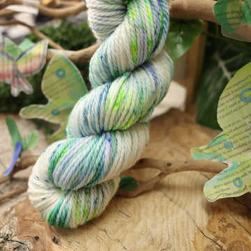 A light blue, teal, green, and white colored Bulky yarn, on top of a plant with bare branches and leaves in the back. along with painted butterflies and leaves made of book pages, all on top of a wooden table.
