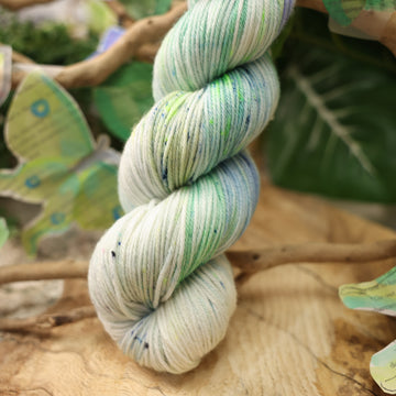 A light blue, teal, green, and white colored DK yarn, on top of a plant with bare branches and leaves in the back. along with painted butterflies and leaves made of book pages, all on top of a wooden table.