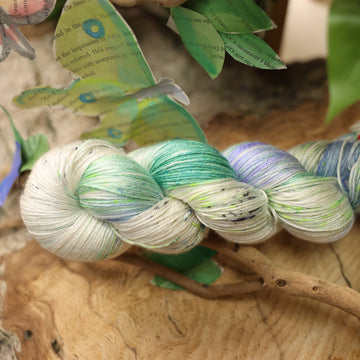 A light blue, teal, green, and white colored silktrue yarn, on top of a plant with bare branches and leaves in the back. along with painted butterflies and leaves made of book pages, all on top of a wooden table.