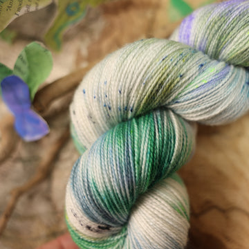 A light blue, teal, green, and white colored sparkle yarn, on top of a plant with bare branches and leaves in the back. along with painted butterflies and leaves made of book pages, all on top of a wooden table.