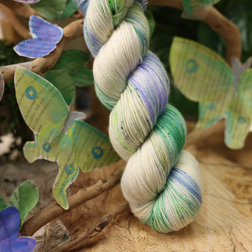 A light blue, teal, green, and white colored sport yarn, on top of a plant with bare branches and leaves in the back. along with painted butterflies and leaves made of book pages, all on top of a wooden table.