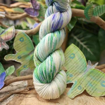 A light blue, teal, green, and white colored worsted yarn, on top of a plant with bare branches and leaves in the back. along with painted butterflies and leaves made of book pages, all on top of a wooden table.
