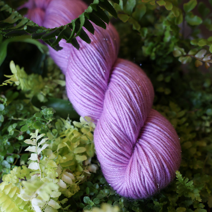 DK sparkle yarn colored a light violet pink color. This skein is on top of a bed of ferns and plants