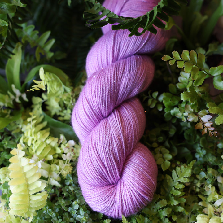 Sparkle fingering yarn colored a light violet pink color. This skein is on top of a bed of ferns and plants