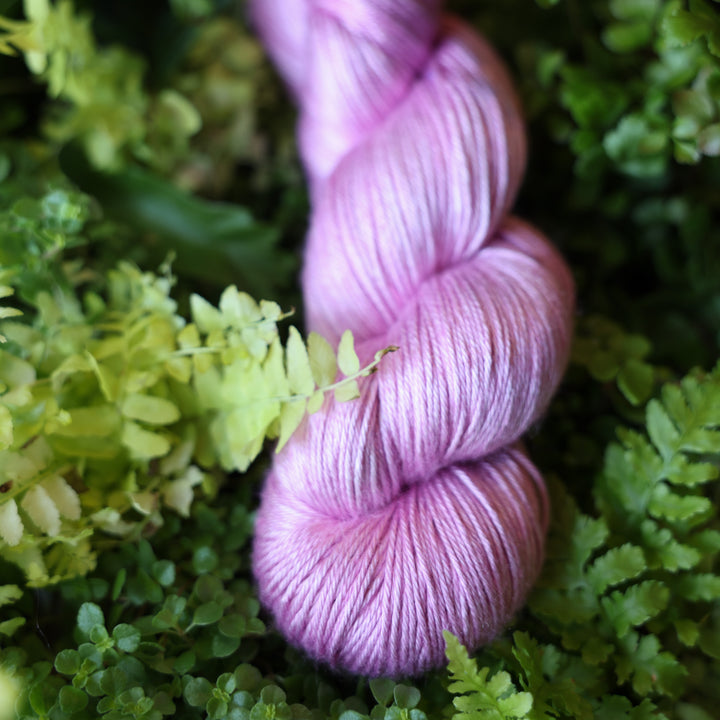 Silk fingering yarn colored a light violet pink color. This skein is on top of a bed of ferns and plants