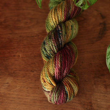 Bulky yarn colored dark shades of green with some reds and yellows mixed in, on top of a brown wooden table and a golden pot in the corner with a plant peaking out.