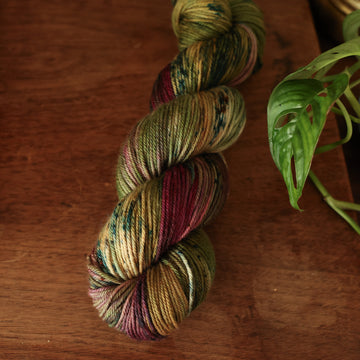 DK yarn colored dark shades of green with some reds and yellows mixed in, on top of a brown wooden table and a golden pot in the corner with a plant peaking out.