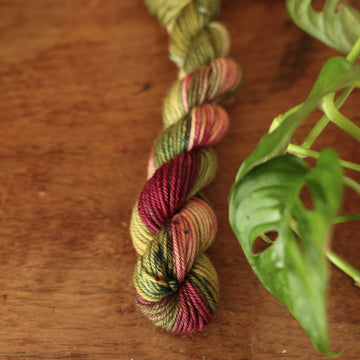 DK mini yarn colored dark shades of green with some reds and yellows mixed in, on top of a brown wooden table and a golden pot in the corner with a plant peaking out.