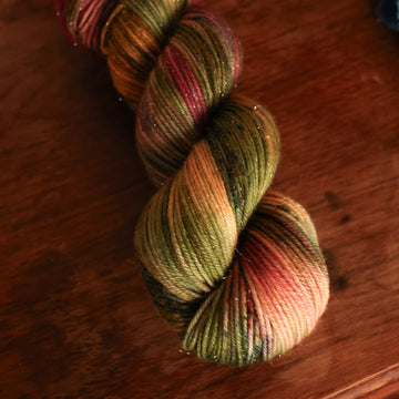 DK Sparkle yarn colored dark shades of green with some reds and yellows mixed in, on top of a brown wooden table and a golden pot in the corner with a plant peaking out.