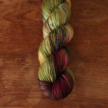 Fingering yarn colored dark shades of green with some reds and yellows mixed in, on top of a brown wooden table.