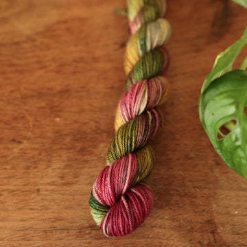 Fingering mini yarn colored dark shades of green with some reds and yellows mixed in, on top of a brown wooden table and a golden pot in the corner with a plant peaking out.