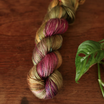 Silk yarn colored dark shades of green with some reds and yellows mixed in, on top of a brown wooden table and a golden pot in the corner with a plant peaking out.