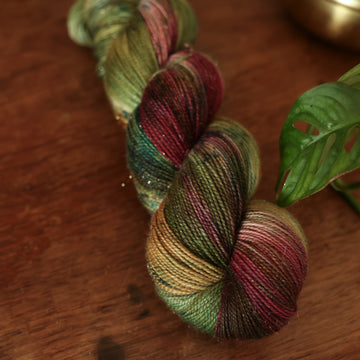 Sparkle Fingering yarn colored dark shades of green with some reds and yellows mixed in, on top of a brown wooden table and a golden pot in the corner with a plant peaking out.