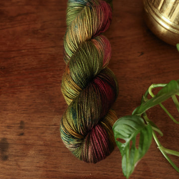 Sport yarn colored dark shades of green with some reds and yellows mixed in, on top of a brown wooden table and a golden pot in the corner with a plant peaking out.