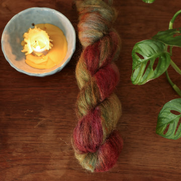 Suri alpaca yarn colored dark shades of green with some reds and yellows mixed in, on top of a brown wooden table and a partially melted in the corner with a plant peaking out.