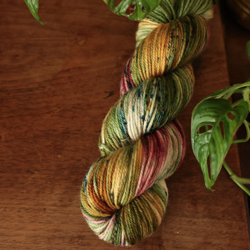 Worsted yarn colored dark shades of green with some reds and yellows mixed in, on top of a brown wooden table and a golden pot in the corner with a plant peaking out.
