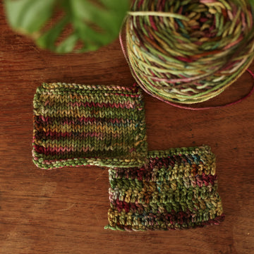 Two knit and crochet swatches colored with warm greens with gentle touches of soft brown, mauve, and blue. Next to the swatches is a caked skein, all sitting on top of a wooden table.