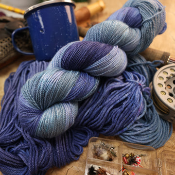 A mixture of different shades of blue fingering yarn next to some fishing equipment like hooks and ends of a pole. and a camping mug in the back along with an unwound version of the yarn. all on top of a wooden table.
