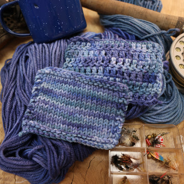 A mixture of different shades of blue crochet and knit swatches next to some fishing equipment like hooks and ends of a pole. and a camping mug in the back along with an unwound version of the yarn. all on top of a wooden table.