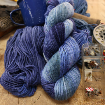 A mixture of different shades of blue worsted yarn next to some fishing equipment like hooks and ends of a pole. and a camping mug in the back along with an unwound version of the yarn. all on top of a wooden table.