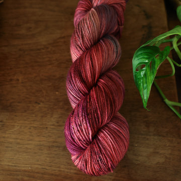 DK yarn colored with different shades of red and some purple and blue colors mixed in, on top of a brown wooden table and a golden pot in the corner with a plant peaking out.