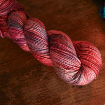 DK sparkle yarn colored with different shades of red and some purple and blue colors mixed in, on top of a brown wooden table and a golden pot in the corner with a plant peaking out.