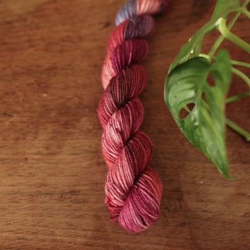 Fingering Mini yarn colored with different shades of red and some purple and blue colors mixed in, on top of a brown wooden table and a golden pot in the corner with a plant peaking out.