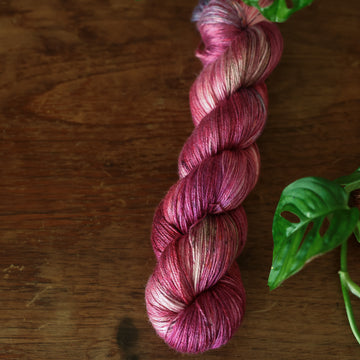 Silk yarn colored with different shades of red and some purple and blue colors mixed in, on top of a brown wooden table  with a plant peaking out.