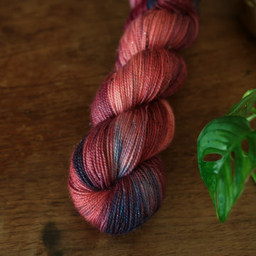 Sparkle Fingering yarn colored with different shades of red and some purple and blue colors mixed in, on top of a brown wooden table and a golden pot in the corner with a plant peaking out.